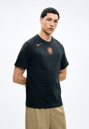 Νεαρός άνδρας με κοντά σγουρά μαλλιά, φορώντας μαύρο μπλουζάκι Nike με πορτοκαλί έμβλημα λιονταριού και μπεζ παντελόνι, στέκεται με τα χέρια πίσω από την πλάτη.