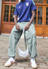 Granatowy sportowy jersey z logo Włoch, jasnoszare spodnie cargo, biała torba na ramię oraz białe sneakersy z czerwonymi paskami. Luźna, outdoorowa sceneria.