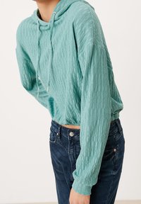 Felpa con cappuccio di un leggero color teal realizzata in un materiale testurizzato, con una vestibilità rilassata e un cordino. Abbinata a jeans blu scuro.
