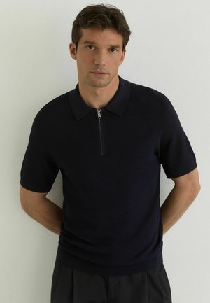 Mees musta lühikeste varrukatega kudumispolo trikooga ja lukuga, seisab tavalise valge seina ees, käed selja taga.