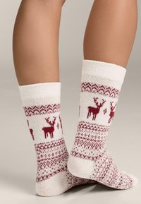 Fehér kötött zokni vörös rénszarvas mintákkal, texturált kialakítással. Puha, meleg anyag, bordázott szegéllyel és mintás díszítéssel.