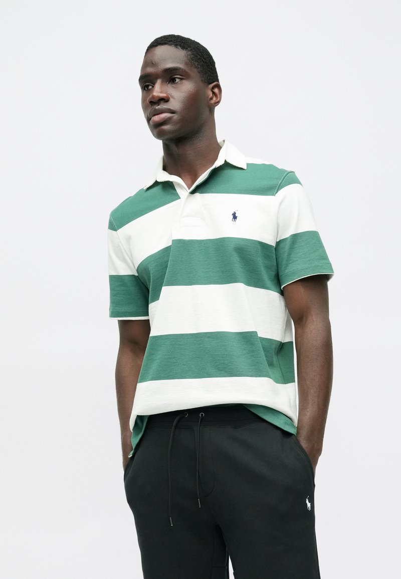 Polo a righe verdi e bianche con colletto, maniche corte e un piccolo logo sul petto, abbinato a pantaloni tuta neri.