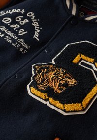 Veste de varsity bleu foncé avec broderie blanche et dorée. Dotée d'un patch de tigre et de détails cousus. Les boutons-pression sont noirs. Texture douce.