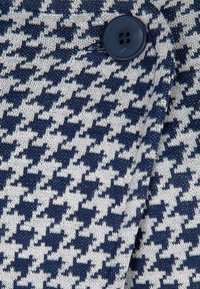 Marinblått och krämfärgat hundtandsmönstrat tyg med en rund marinblå knappdetalj. Texturen ser stickad och något texturerad ut.