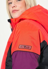 Vaatteet, joissa on kirkkaan oranzhi, musta ja lilla väriyhdistelmä. Merkkijacketissa on huppu, joustavat hihansuut ja suuri etiketti hihassa.