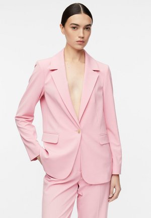 Kobieta z zaczesanymi do tyłu ciemnymi włosami, ubrana w jasno-różową, dopasowaną marynarkę i pasujące spodnie, ręka w kieszeni, neutralny wyraz twarzy.
