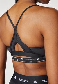 Svart sportsbh med racerback-design og utskåret detalj. Har en merket elastisk band med hvit tekst. Glatt, elastisk stoff.