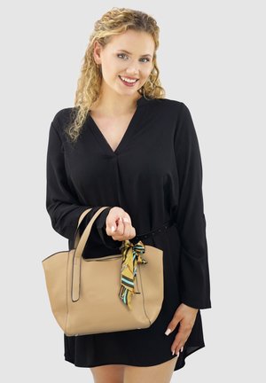 Beige leren tote bag met gestructureerde vorm, voorzien van bovenhandvatten en een patroonsjaal die aan het handvat is geknoopt. Zwart langemouwen jurk.