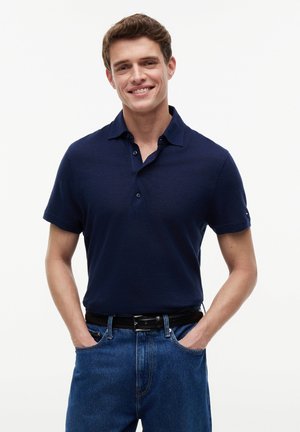 Usmívající se muž v tmavě modrém tričku s krátkým rukávem, zastrčeném do modrých džínů, s rukama v kapsách na bílém pozadí.