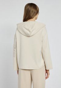 Beżowa bluza z kapturem o lu�źnym kroju, długimi rękawami i boczną kieszenią na lewym rękawie. Gładka faktura i minimalistyczny design.
