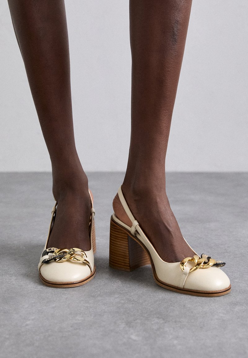 Dunkelhäutige Beine, die beige Leder-Slingback-High Heels mit Holzblockabsätzen und Goldkettendetails auf einem grauen Boden tragen.