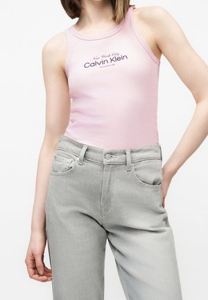 Nainen pukeutunut vaaleanpunaiseen Calvin Kleinin hihattomaan toppiin ja vaaleanharmaisiin korkeavyötäröisiin farkkuihin, käsi taskussa.