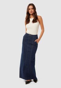 Fustă maxi din denim cu croială dreaptă, de culoare albastru închis, cu cusături vizibile și două buzunare laterale; asortată cu un top alb fără mânecă.