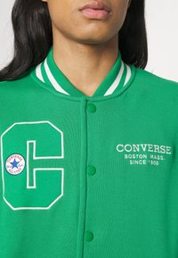 Chaqueta varsity de algodón verde con cuello de rayas blancas, logo 'C' bordado y texto de Converse. Presenta botones a presión y una textura suave.