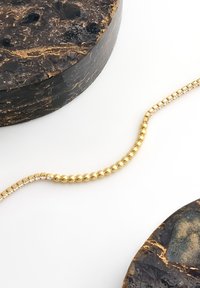 Guldbelagd armband med små fyrkantiga kristaller placerade på en vit yta bredvid två runda svarta och guldtexturerade stenar.