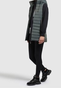 Gilet imbottito verde con cappuccio, dotato di zip frontale e dettagli neri. Indossato sopra una maglia nera a maniche lunghe e leggings neri, abbinati a sneakers nere robuste.