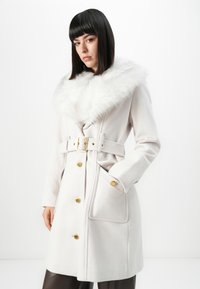 Vrouw met steil, donker haar, gekleed in een witte jas met riem, grote bontkraag en gouden knopen, staande tegen een witte achtergrond.