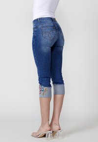 Jean capri en denim bleu avec des détails floraux brodés sur l'ourlet. Lavage délavé, design ajusté, avec poches arrière et poignets effilochés.