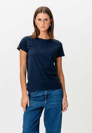 Kvinde står op, iført navyblå kortærmet skjorte og blå jeans, med glat brunt hår og blomstertatovering på venstre overarm.