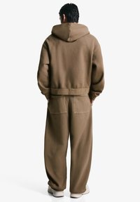 Brun oversized hoodie med en lomme og snoretræk i huen, parret med matchende løstsiddende bukser, placeret mod en hvid baggrund.