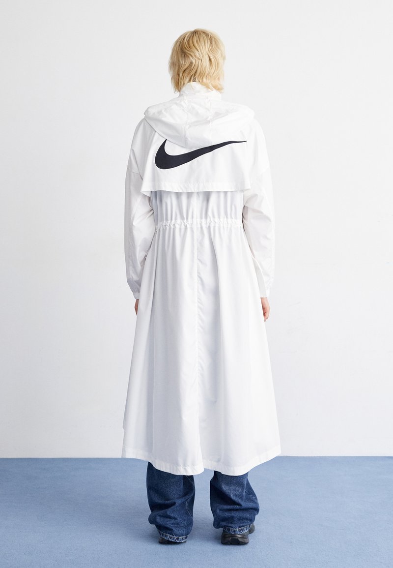 Lange witte windbreaker met capuchon, elastische taille, zwart Nike-logo op de achterkant; heeft lange mouwen en gaat tot halverwege de kuit.