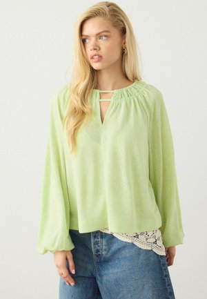 Blondynka ubrana w luźny, jasnozielony top z długim rękawem z marszczonym dekoltem i dwoma poziomymi paskami, zestawiona z niebieskimi dżinsami i koronkową lamówką.