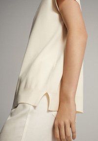 Kermaisen värinen lyhythihainen paita neulotusta kankaasta, jossa on kaareva helma ja sivuhalkiot liikkeen helpottamiseksi, sileällä pinnalla.