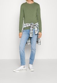 Groene lange mouwen shirt, lichtblauwe jeans, witte sneakers en een geruit shirt om de taille gebonden. Casual en ontspannen pasvorm.