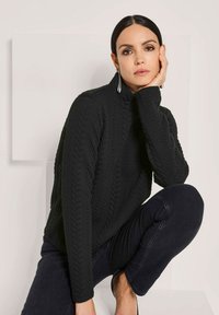 Schwarzer Strickpullover mit Rollkragen und langen Ärmeln, der ein strukturiertes Muster und gerippte Bündchen aufweist. Getragen mit dunklen Jeans.