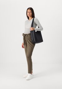 Sac fourre-tout en cuir noir avec bandoulière réglable, surface lisse, design minimaliste. Le modèle porte un haut à manches longues gris et un pantalon vert olive.