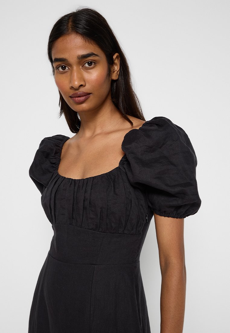 Jeune femme aux longs cheveux noirs portant une robe noire avec des manches courtes bouffantes et un décolleté carré froncé, regardant en avant.