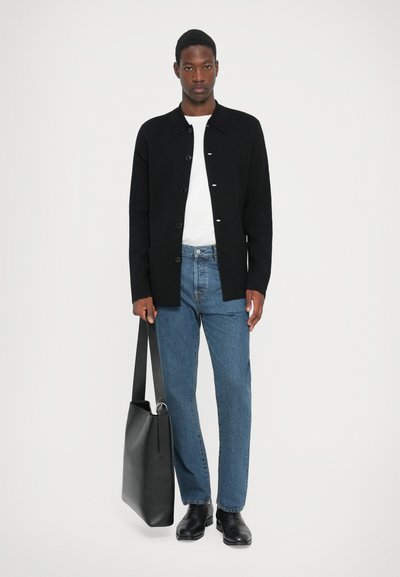 Zwarte button-up jas, witte T-shirt, blauwe spijkerbroek, zwarte schoenen en een zwarte tote bag. De jas heeft een klassieke kraag en zijzakken.