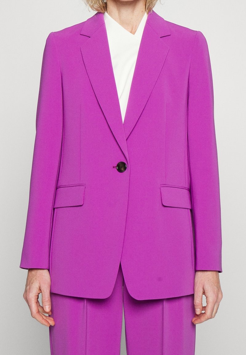 Femme portant un blazer violet vif cintré avec un unique bouton noir et un pantalon assorti, associé à un haut blanc en dessous.