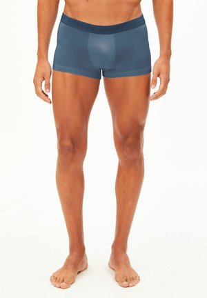 Modello maschile in piedi a piedi nudi indossando boxer blu su uno sfondo bianco semplice, visibile dalla vita ai piedi.