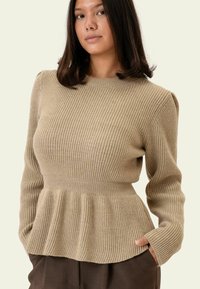 Beiger Rippstrickpullover mit tailliertem Schnitt, ausgestelltem Saum und langen Ärmeln. Die Textur ist weich und zeigt dezente vertikale Linien sowie Raffungen an der Taille.