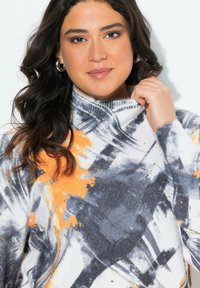 Frau mit welligem, dunklem Haar, die silberne Creolen trägt und einen Pullover mit abstraktem Muster in Weiß, Orange und Grau hält und den Kragen fasst.