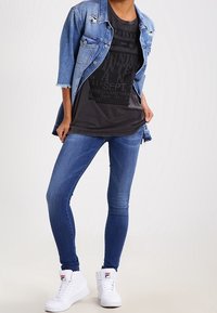 Denim jacket with distressed details over a black graphic t-shirt, paired with blue skinny jeans and white high-top sneakers.