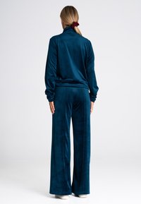 Turkusowy dres z weluru z wysokim kołnierzem, długimi rękawami i szerokimi spodniami. Gładka tekstura z delikatnym połyskiem, luźny krój.