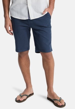Mann trägt marineblaue Shorts, weißes Hemd und schwarze Flip-Flops mit grünen Riemen, steht mit einer Hand in der Tasche vor weißem Hintergrund.