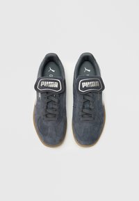 Mėlyni zomšiniai sportbačiai su pilkais raišteliais ir guminėmis padais, ant liežuvėlio ir šono matomas ryškus sidabrinis Puma logotipas.