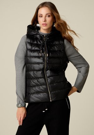 Donna con i capelli lunghi che indossa un gilet imbottito nero con cappuccio sopra una maglia grigia a maniche lunghe e pantaloni neri, in piedi con una mano sul fianco.