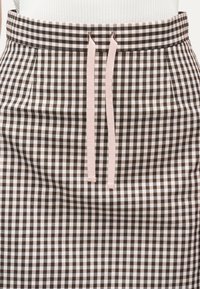 Περιφέρεια καφέ και λευκής φουστας με καρό σχέδιο, με ροζ κορδόνι δεμένο μπροστά και λευκό ριμπ τοπ που έχει φορεθεί μέσα στη φούστα.