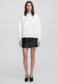 Sudadera blanca con bordado floral, combinada con una mini falda de cuero sintético negra. Tacones negros de punta completan el conjunto.