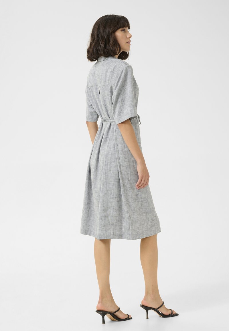 Mujer con cabello oscuro a la altura de los hombros, vestida con un vestido gris hasta la rodilla con cinturón y sandalias negras de tacón, de pie sobre un fondo blanco.