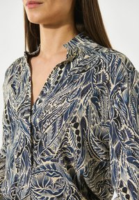 Camisa de cuello hecha de una tela ligera, con un patrón floral en azul y negro sobre un fondo beige claro, vista en primer plano.