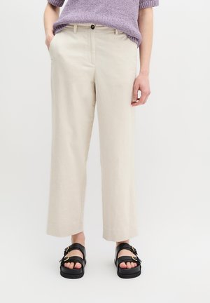 Personne portant un pantalon large crème, des sandales noires à boucle et un pull tricoté violet à manches courtes, debout avec une main dans la poche.