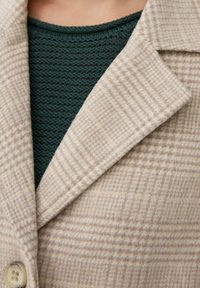 Beige en crème geruite wollen jas met revers over een donkergroene gebreide sweater op de bovenste helft van een persoon.