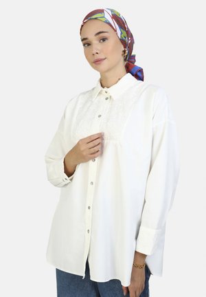 White button-up shirt with embroidered front panel, long sleeves, and button cuffs. Model wears a colorful patterned headscarf.