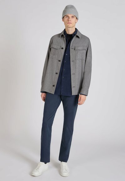 Grijze wollen overhemdjas met vier zakken aan de voorkant, gecombineerd met een marineblauw overhemd en marineblauwe broek. Witte sneakers maken de outfit compleet.