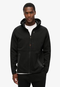 Zwarte zip-up hoodie met een hoge kraag, zijzakken met rits en een gladde textuur. Bevat subtiele logo-details op de borst.